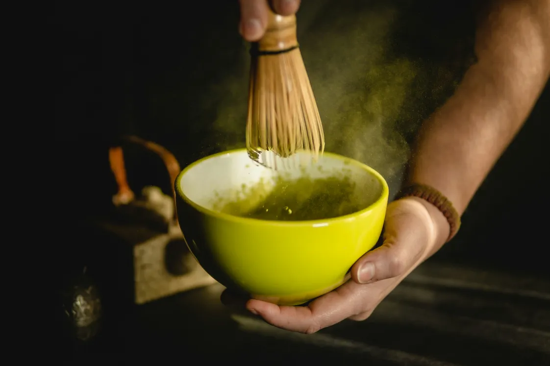 a close-up photograph of someone mixing matcha using a traditional 'Chasen' (bamboo whisk) in a green bowl