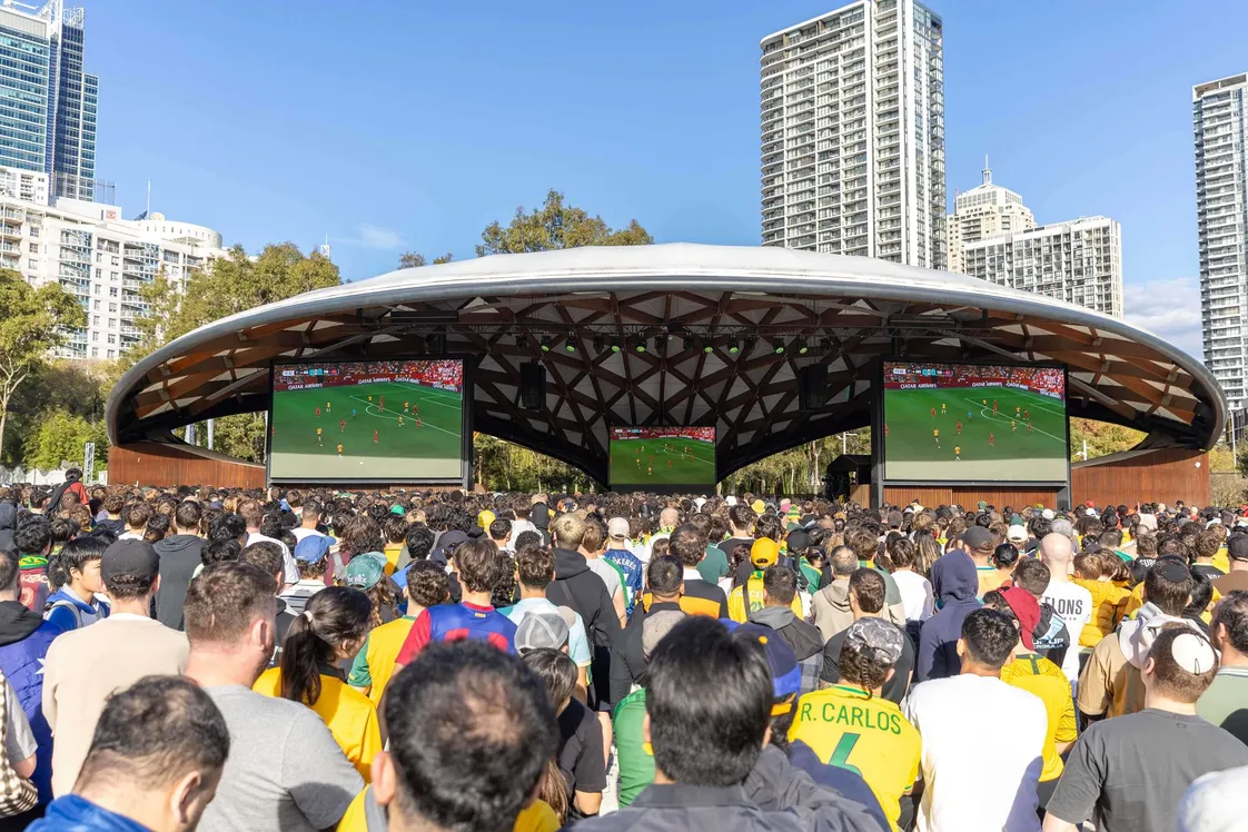 Fans wearing yellow and green jerseys watching soccer at the 2026 World Cup fan zone at Tumbalong Park in Darling Harbour on a sunny day